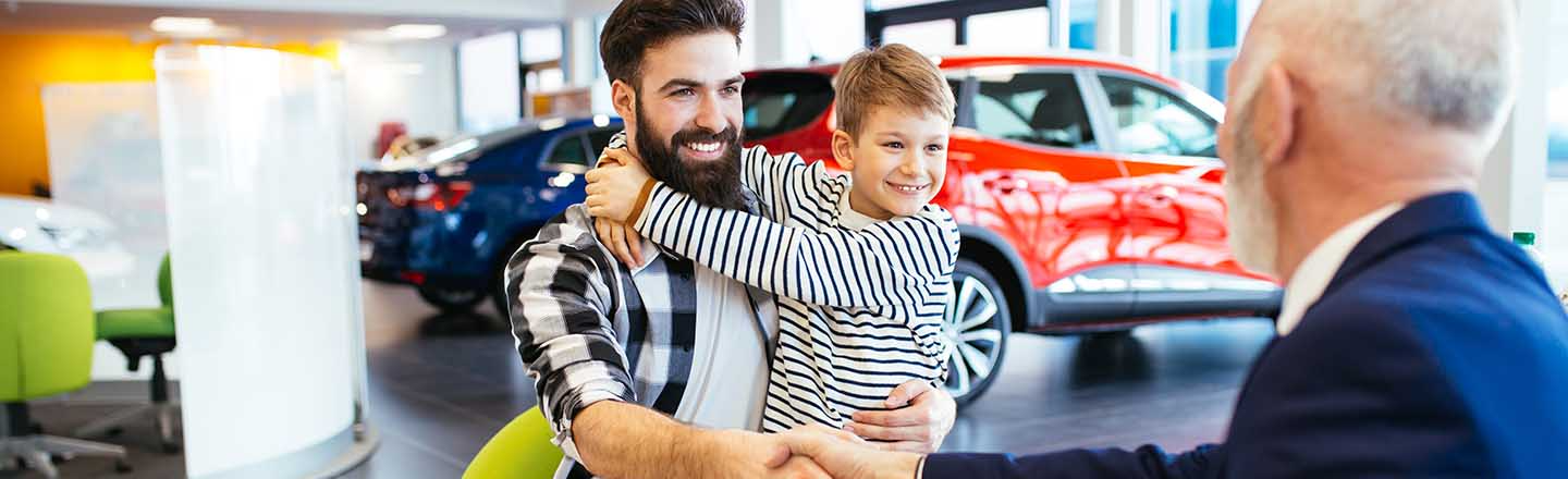 A smiling father holding his son and shaking hands with a dealership sales representative