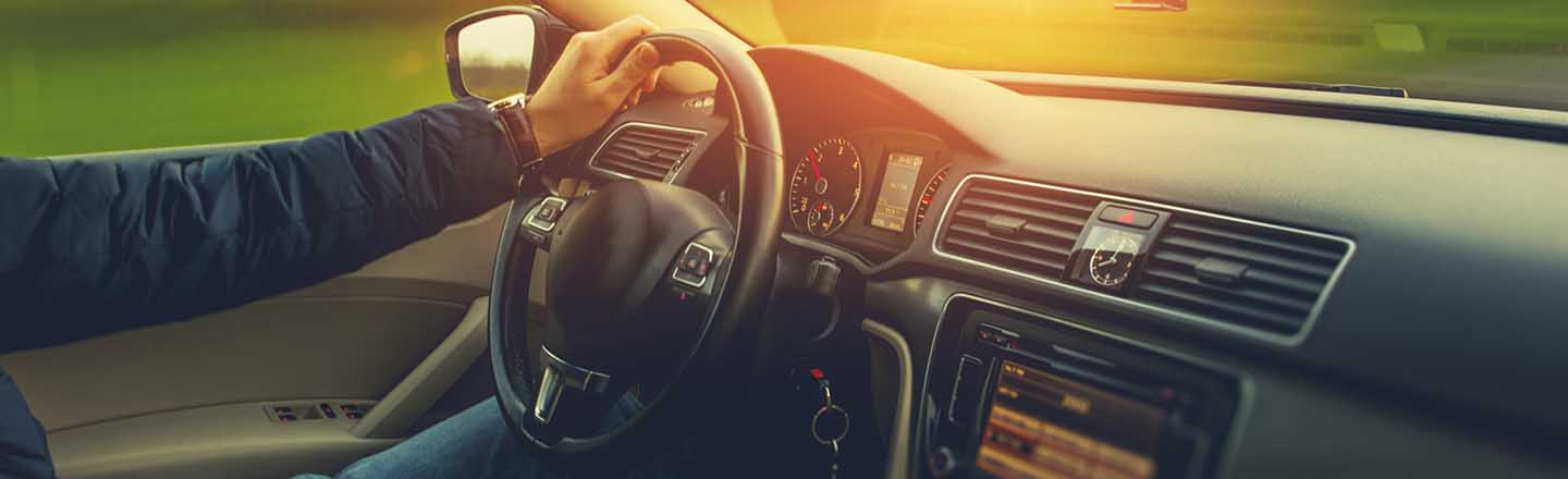 A close up of a single hand on the wheel of a car while it's driving, with greenery and a light flare outside